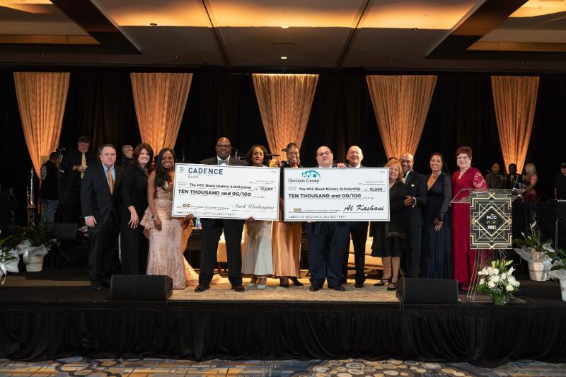 a photo displaying men and women lined across the stage and two are holding two big check images